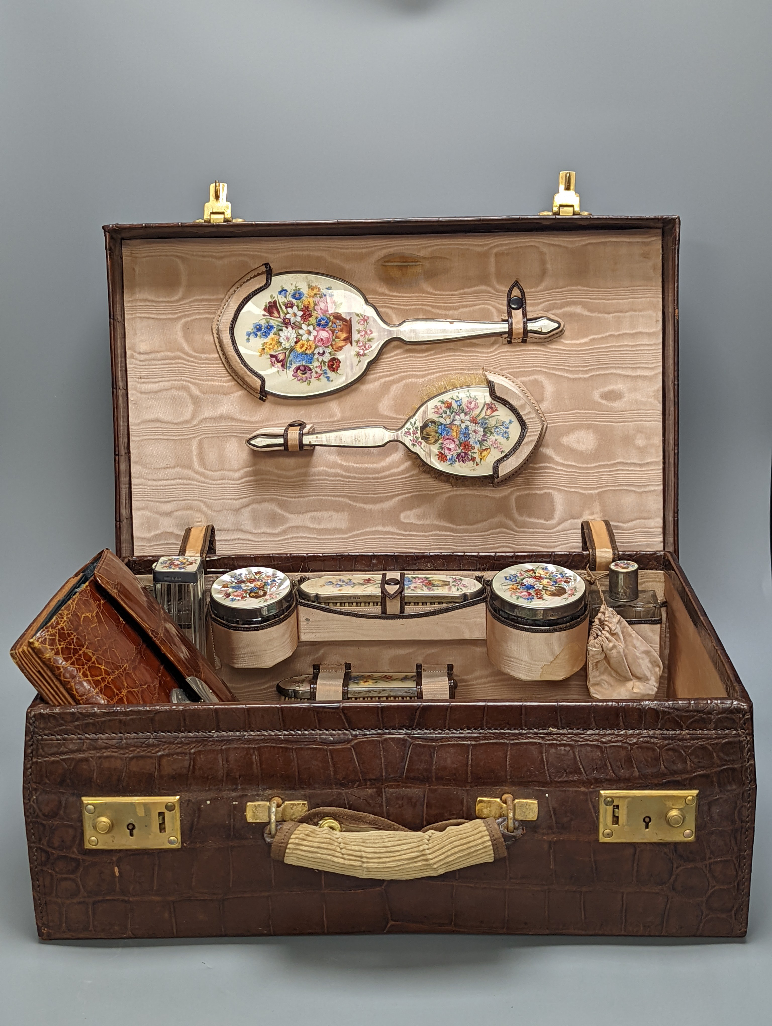 A 1930's crocodile skin travelling toilet case, with ten silver and floral enamel mounted accoutrements, Henry Clifford Davis, Birmingham, 1935,6, together with five other silver and enamel brushes and mirror etc and a c